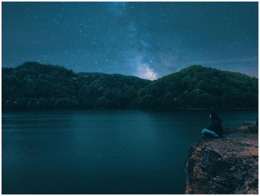 A person sits on a cliff overlooking a serene lake