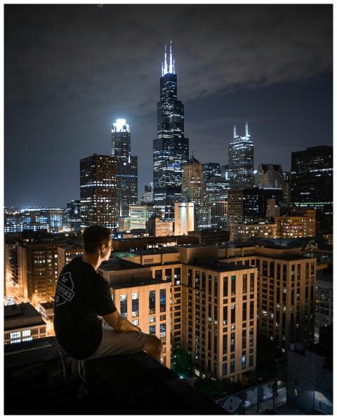 A man sits on a rooftop, overlooking the illuminat