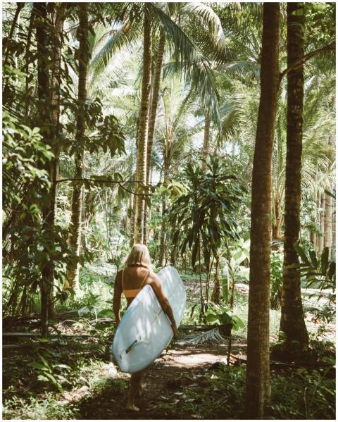 A young woman carries a surfboard through lush, su