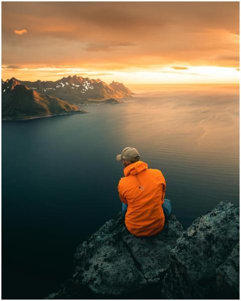 Man in orange jacket sits on cliff, overlooking a