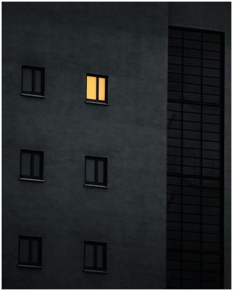 A dark building facade with one window lit, creati