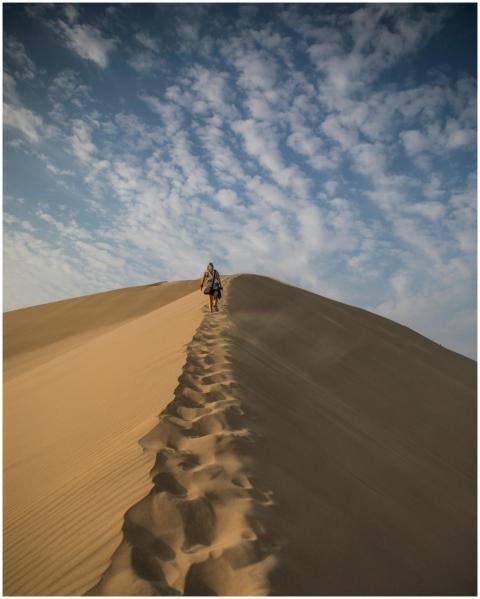 A person walks along the sand dunes under a blue s