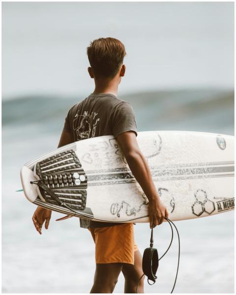 Back view of a young surfer carrying a board by th