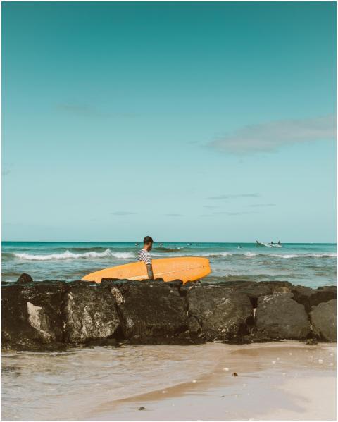 A lone surfer stands on a rocky beach with a surfb