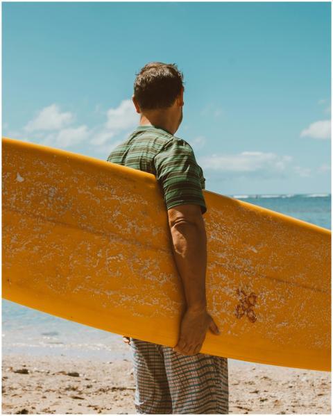 A man stands on a sunny beach holding a yellow sur