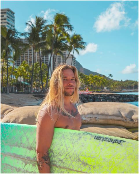 A tattooed man with long hair holding a surfboard