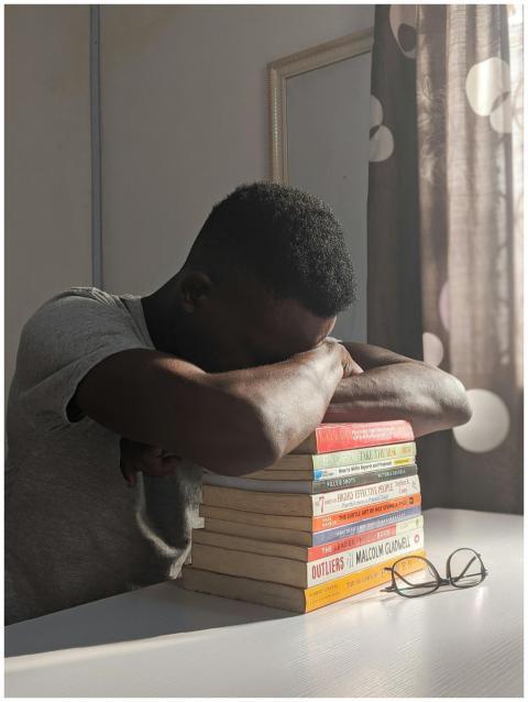 A young man rests on a stack of books at a desk, s