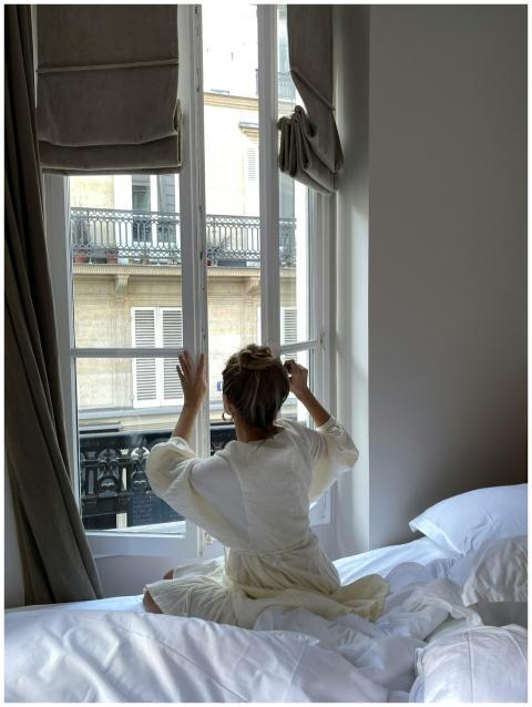 A woman sitting on a bed opens a window in a Paris