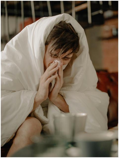 A young man wrapped in a blanket sneezes, indicati