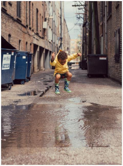 A young boy in a yellow outfit jumps over a puddle
