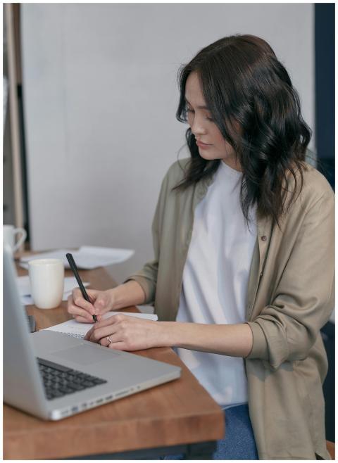 Adult woman writing notes while using a laptop at