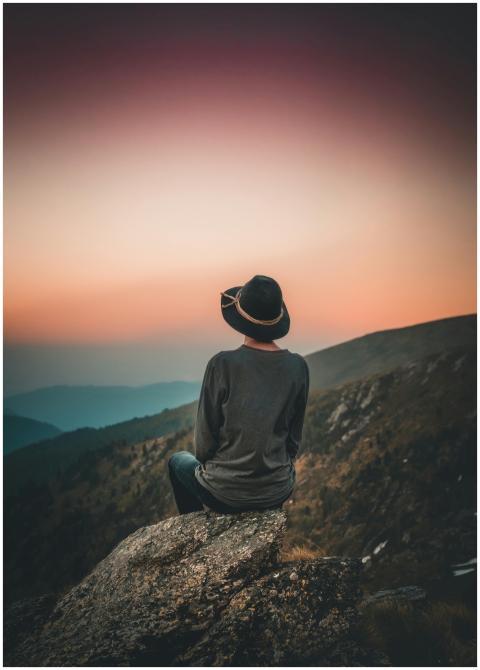 Person in hat sits on rock, gazing at sunset over