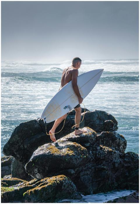 Surfer holding a surfboard stands on a rocky beach
