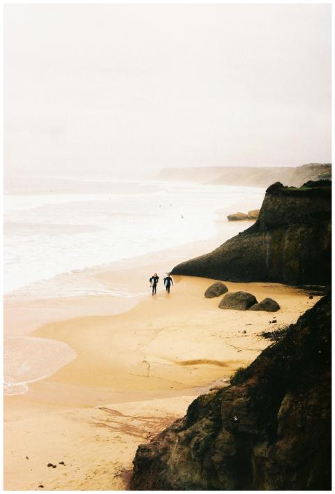 Two surfers walk along a misty beach with towering