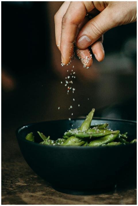 A hand sprinkles salt on fresh edamame, showcasing