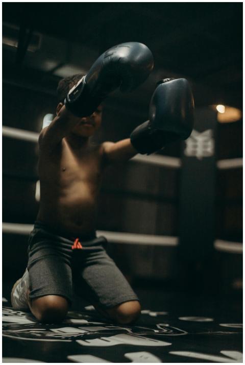A young boy kneels in a boxing ring with gloves, s
