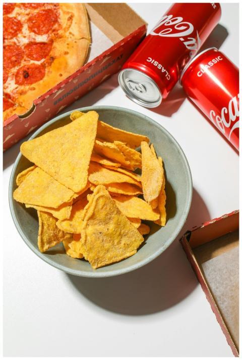 A bowl of tortilla chips with pizza and cola cans,