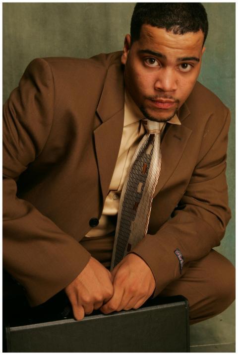 A confident man in a brown suit sits holding a bri