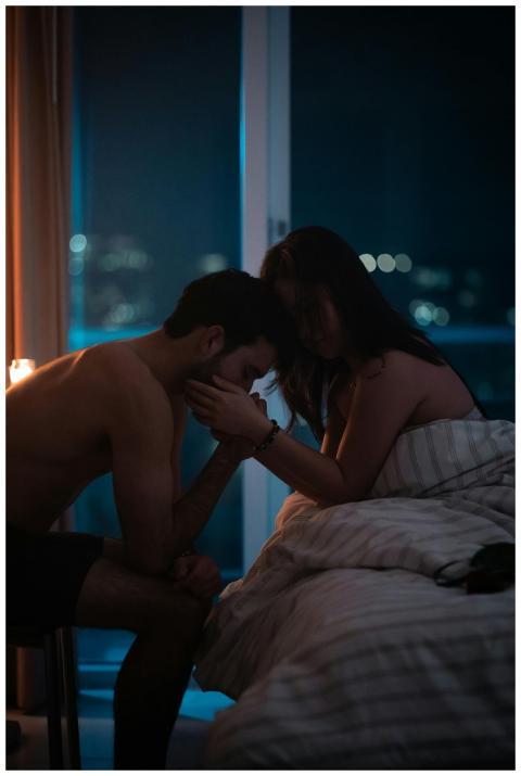 A couple shares a tender moment in a dimly lit bed