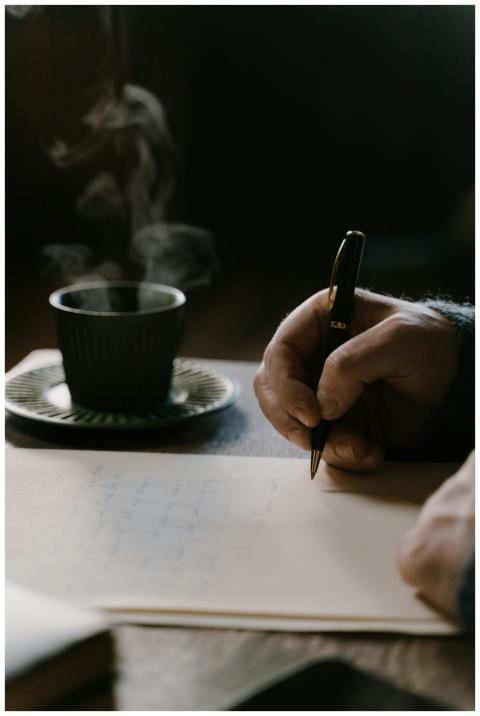 A hand holds a pen over paper with a steaming cup