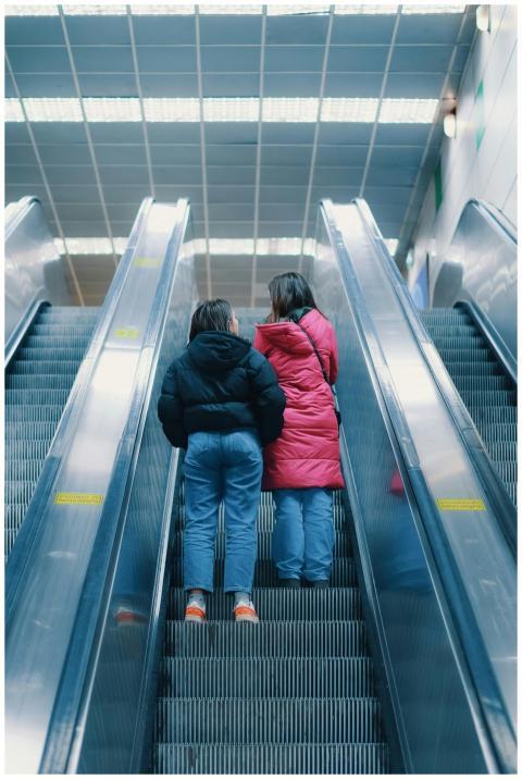 Two women in puffer jackets stand on escalators in