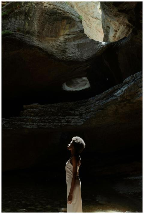 A woman stands thoughtfully inside a cave, illumin