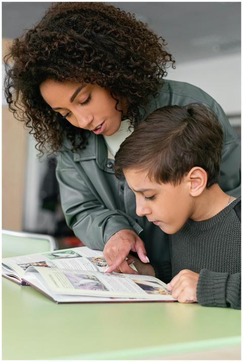 A woman helps a boy read in a classroom setting. E