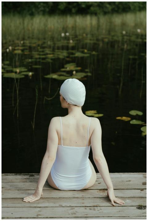 Woman in swimsuit and cap sitting on dock by a ser