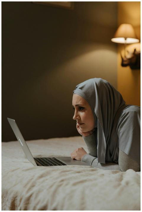 A Muslim woman in a hijab lies on a bed using a la