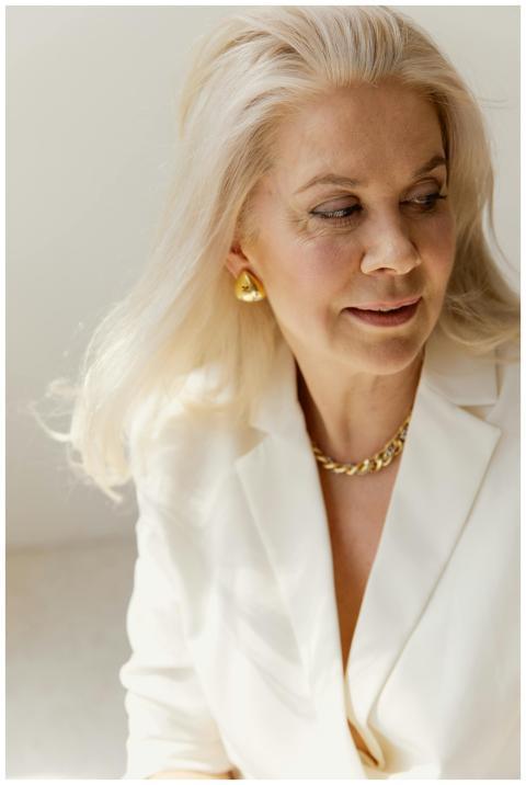 A graceful senior woman in a white blazer with gol