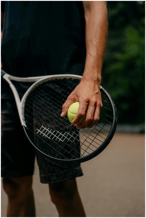 A hand gripping a tennis ball and racket outdoors,
