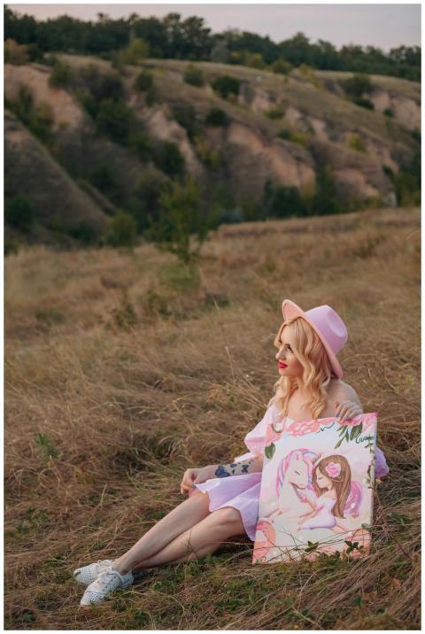 A woman in a pink dress and hat sits with a painti