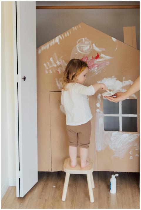 Child painting a cardboard house indoors with crea