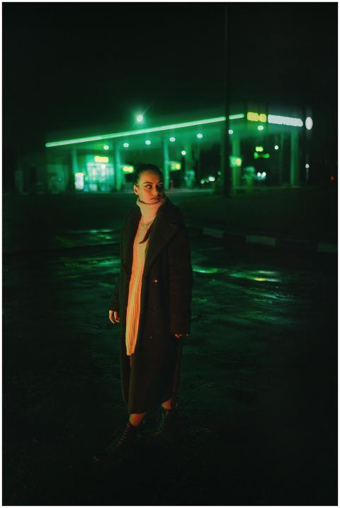 A woman stands near a neon-lit gas station at nigh