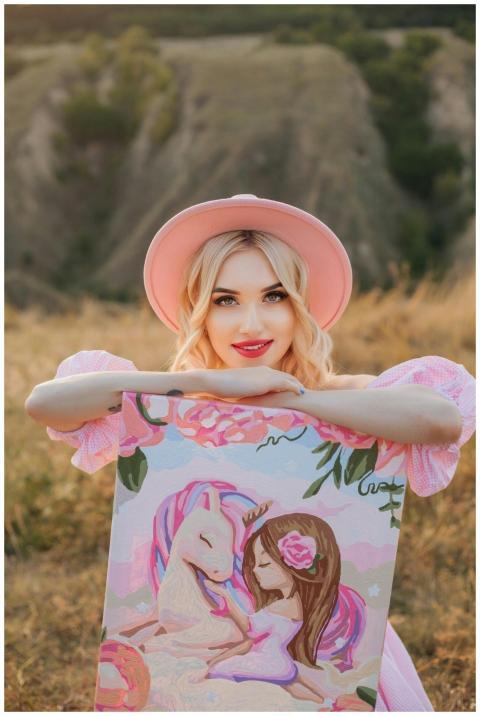 A woman in a pink hat poses with a unicorn paintin