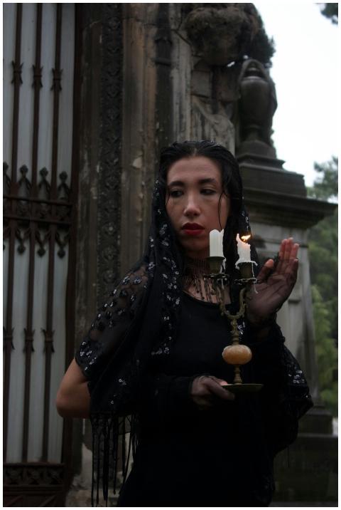 A woman in gothic attire holds a candle outside an