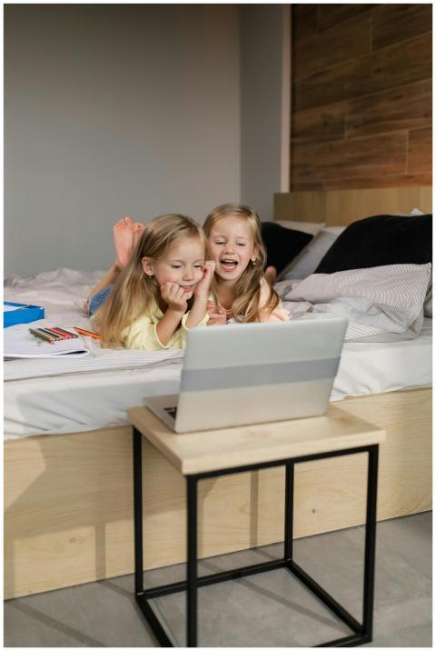 Two joyful twin girls learning online, lying on be