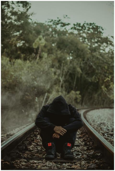 A person in a hoodie sits on empty train tracks, s