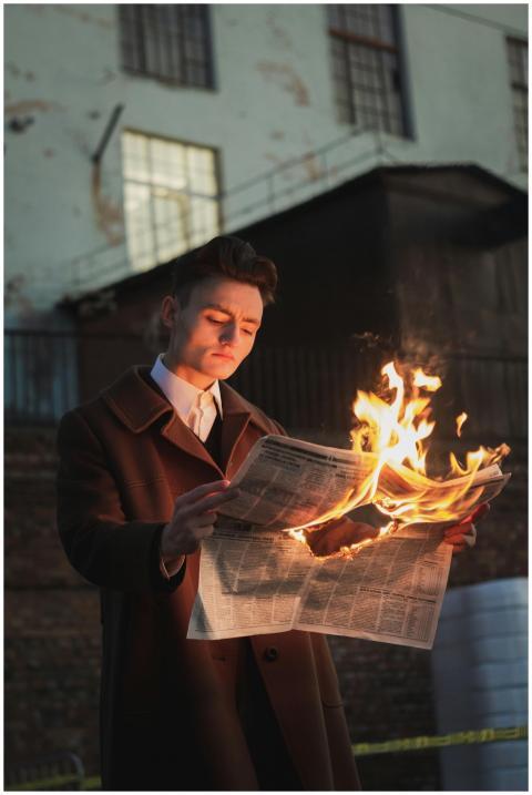 A man in a coat reads a burning newspaper outside
