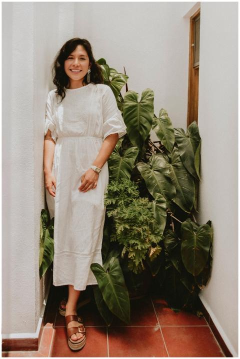 A smiling woman in a white dress stands indoors su