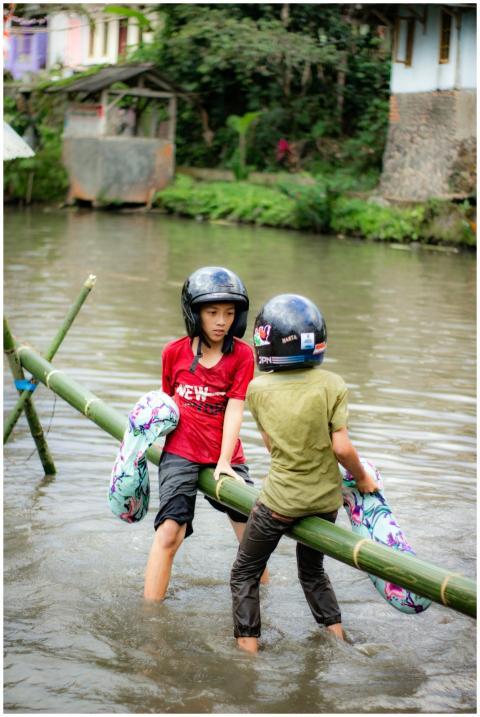 Two kids with helmets balance on bamboo over a riv