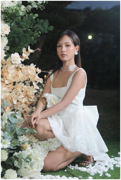 A young woman in a white dress kneels by a flower
