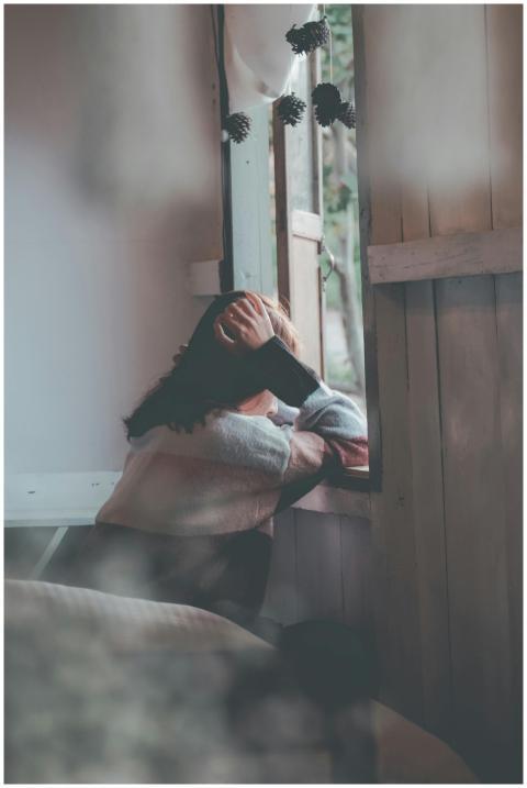 A woman sits indoors gazing thoughtfully out of a