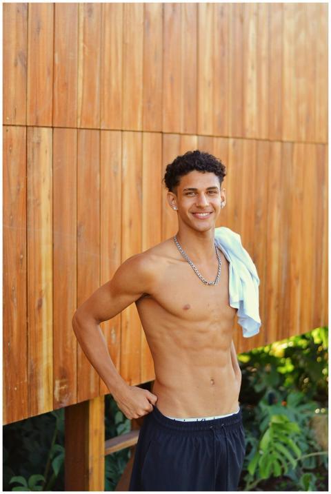 A smiling shirtless man stands outdoors by a woode