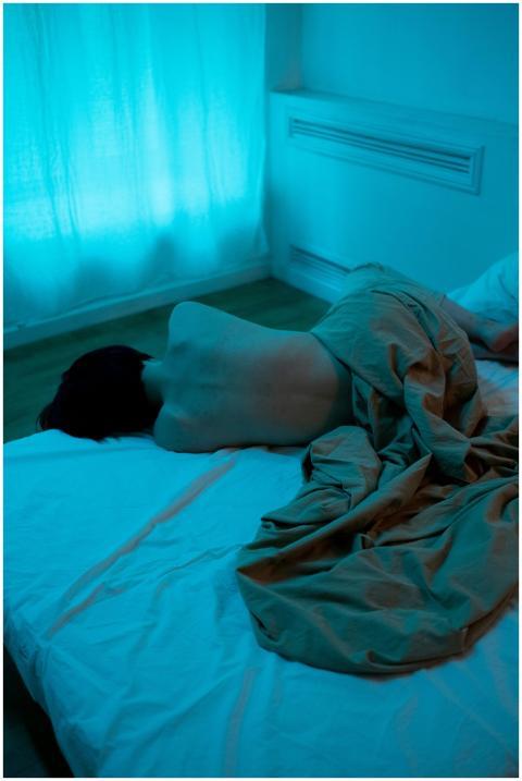 A person lies on a bed in a dimly lit, moody room,
