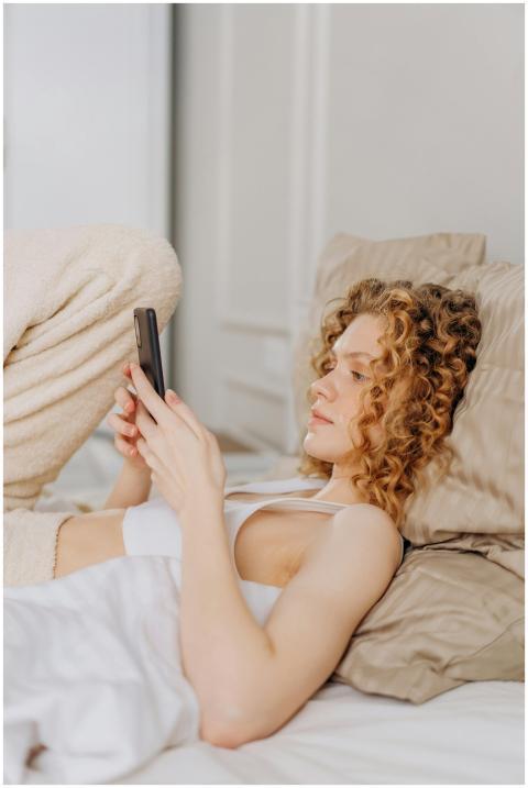 Young woman with curly hair lying in bed, using sm