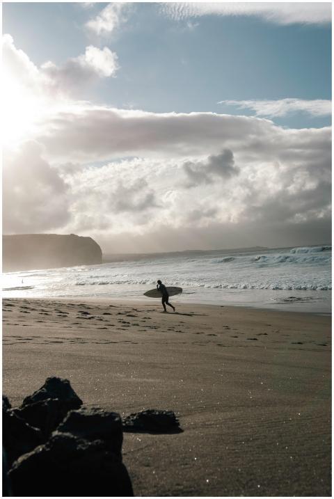 A lone surfer walks along the sandy beach against