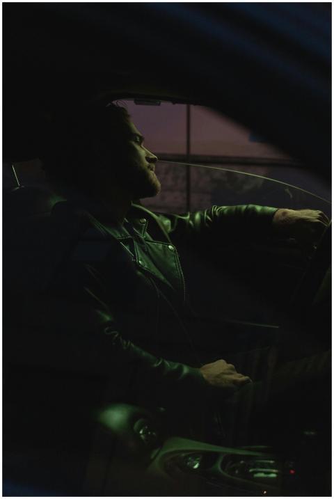 A man in a leather jacket sits in a car, driving a