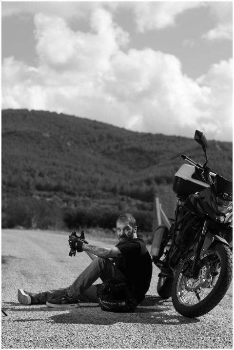 A man sits beside his motorcycle on a rural road,