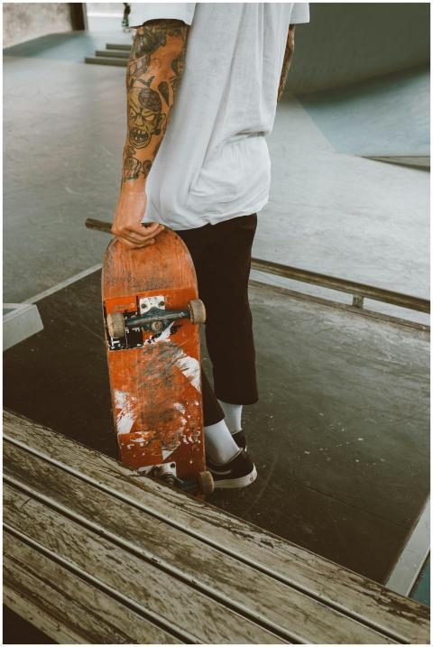 A tattooed skateboarder stands holding a skateboar